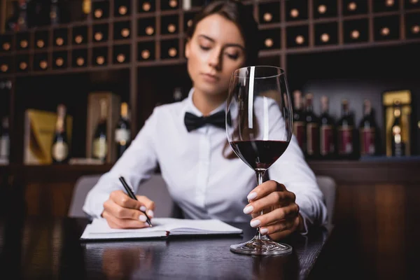 Foco seletivo de sommelier segurando copo de vinho enquanto escreve no caderno à mesa — Fotografia de Stock