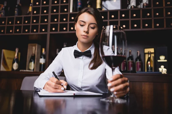 Enfoque selectivo de la escritura joven sommelier en el cuaderno cerca de la copa de vino durante la degustación - foto de stock