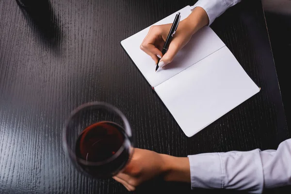 Обрізаний вид сомельє, що пише на блокноті, тримаючи келих вина за столом — стокове фото