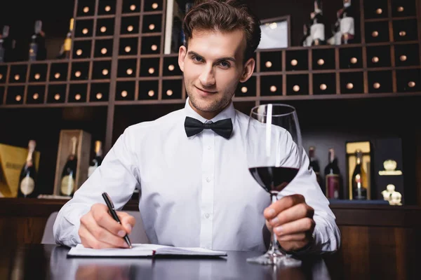 Selektiver Fokus des Sommeliers, der in die Kamera blickt, während er im Restaurant ein Glas Wein hält und auf ein Notizbuch schreibt — Stockfoto