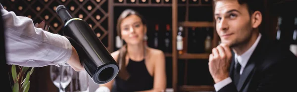 Cultura horizontal de sommelier segurando garrafa de vinho perto de casal elegante no restaurante — Fotografia de Stock
