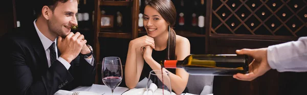 Panoramic shot of young couple sitting near sommelier pouring wine in restaurant — Stock Photo