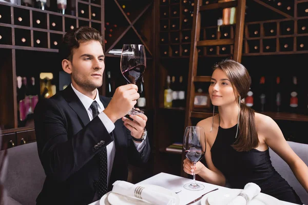 Joven de traje mirando una copa de vino cerca de su novia en el restaurante - foto de stock