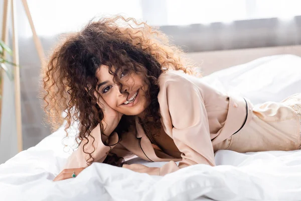 Joyful and curly woman looking at camera while lying on bed — Stock Photo