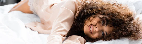 Panoramic crop of curly woman looking at camera while lying on bed — Stock Photo