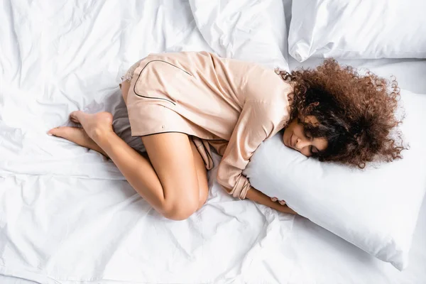 Ansicht von oben: lockige Frau mit geschlossenen Augen, die sich im Bett an ein Kissen schmiegt — Stockfoto