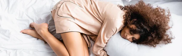 Vista superior da mulher encaracolada com olhos fechados abraçando travesseiro enquanto dorme na cama, conceito panorâmico — Fotografia de Stock