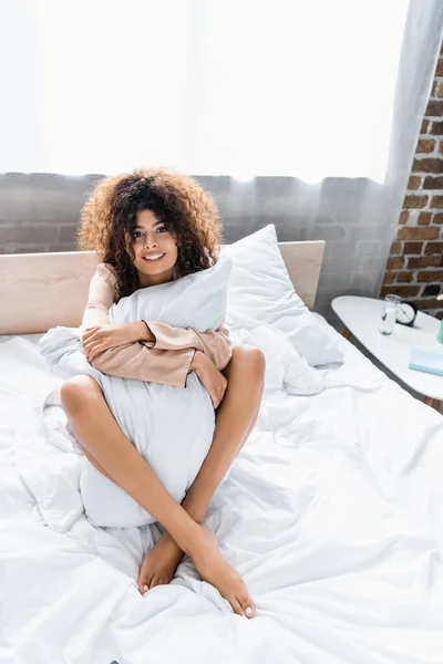 Довольная и босая женщина, сидящая с подушкой на кровати — стоковое фото