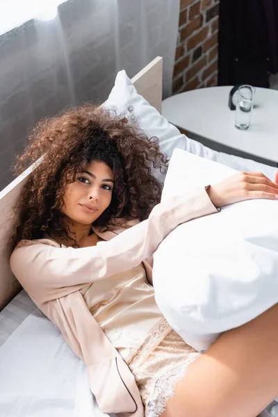 Vue grand angle de jeune femme bouclée couchée sur le lit et tenant oreiller — Photo de stock