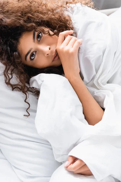 Femme bouclée enveloppée dans une couverture blanche regardant la caméra — Photo de stock