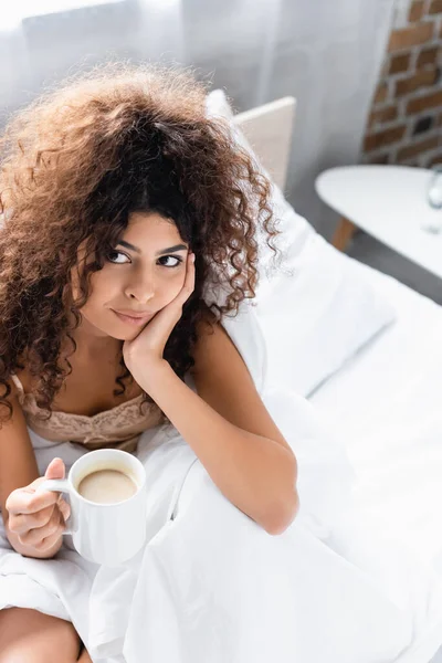 Vista de ángulo alto de la mujer joven rizada sosteniendo la taza de café en la cama - foto de stock