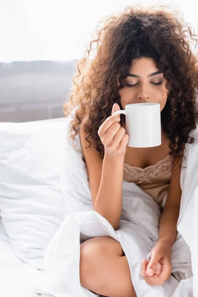 Giovane donna riccia con gli occhi chiusi odore di caffè in camera da letto — Foto stock