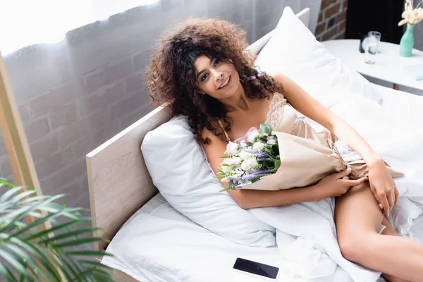 Foyer sélectif de la femme bouclée avec des fleurs regardant la caméra près du smartphone avec écran vide — Photo de stock