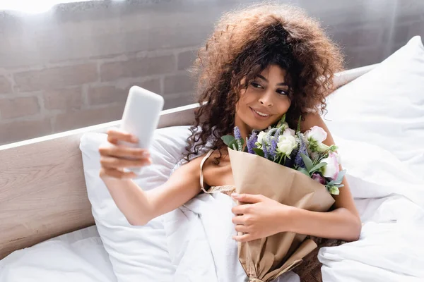 Enfoque selectivo de la mujer complacida tomando selfie con flores en el dormitorio - foto de stock