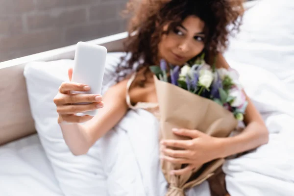 Selektiver Fokus der lockigen Frau beim Selfie mit Blumen im Schlafzimmer — Stockfoto