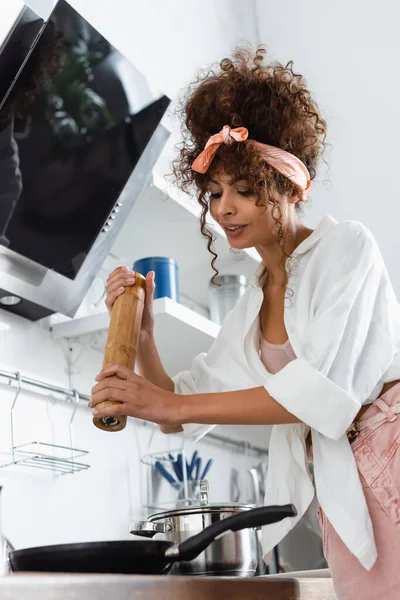 Curly woman holding salt mill above frying pan in kitchen — Stock Photo