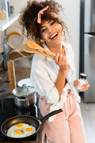 Donna gioiosa che tiene pinze da cucina e bottiglia con sale vicino alle uova sulla padella — Foto stock