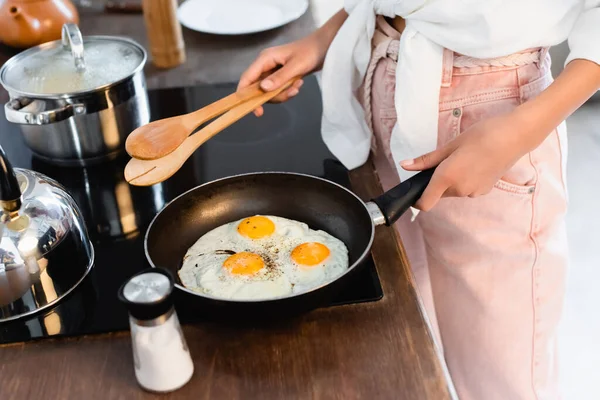 Cropped view of woman holding kitchen tongs near eggs on frying pan — Stock Photo