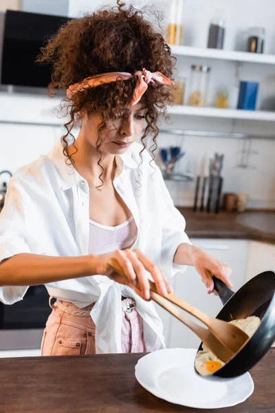 Mujer joven rizada sosteniendo sartén y pinzas de cocina mientras sirve huevos fritos en el plato - foto de stock
