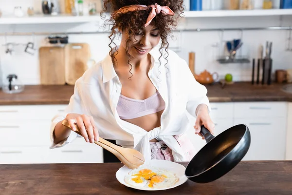 Mujer alegre sosteniendo sartén y pinzas de cocina mientras sirve huevos fritos en el plato - foto de stock