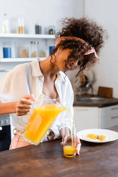 Joyful woman holding jug and pouring fresh orange juice near glass and plate with fried eggs — Stock Photo