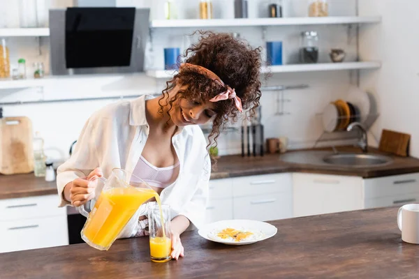 Mulher encaracolado e alegre segurando jarro e derramando suco de laranja fresco perto de vidro e prato com ovos fritos — Fotografia de Stock