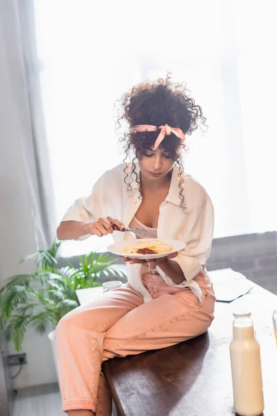 Curly woman sitting on table and holding fork near plate with prepared eggs — Stock Photo