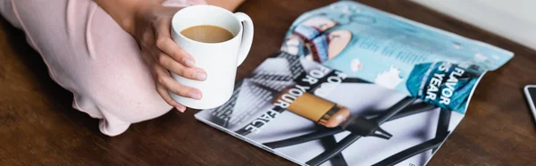 Horizontal crop of woman holding cup of coffee near magazine on table — Stock Photo