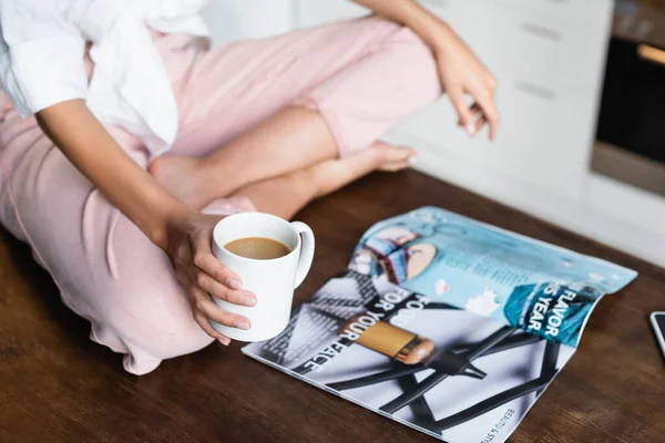 Обрезанный вид женщины, держащей чашку кофе возле журнала на столе — стоковое фото