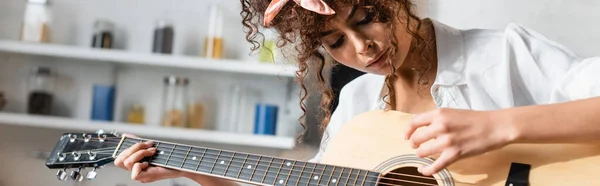 Panoramic crop of curly woman playing acoustic guitar at home — Stock Photo