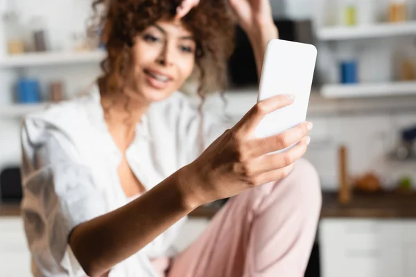 Selective focus of curly woman taking selfie on smartphone in kitchen — Stock Photo