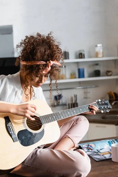 Giovane donna riccia che suona la chitarra acustica vicino alla tazza sul tavolo — Foto stock