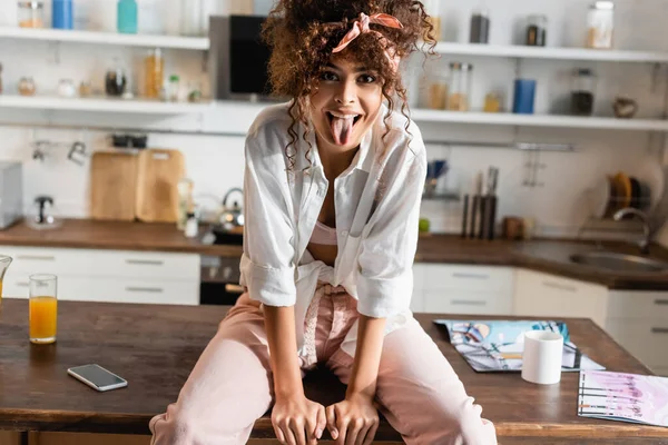 Femme bouclée assise sur la table et sortant la langue — Photo de stock