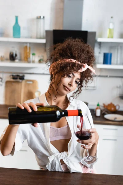 Curly woman holding bottle and pouring red wine in glass — Stock Photo