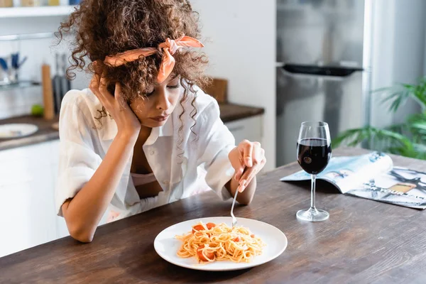 Mujer molesta sosteniendo tenedor cerca del plato con espaguetis preparados y copa de vino en la mesa - foto de stock