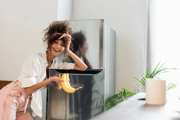 Selective focus of excited woman holding bananas near fridge in kitchen — Stock Photo