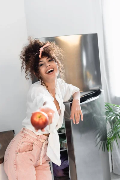 Selective focus of joyful woman holding apple near fridge in kitchen — Stock Photo