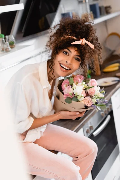 Focalizzazione selettiva di gioiosa giovane donna che tiene fiori in cucina — Foto stock