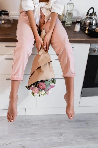 Cropped view of woman sitting on wooden surface with flowers in kitchen — Stock Photo