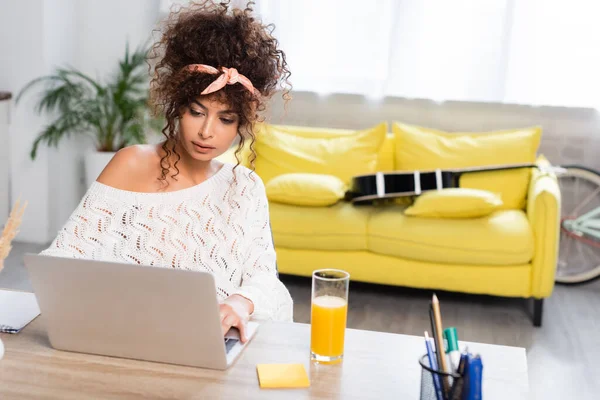 Freelancer rizado mirando portátil cerca de vaso de jugo de naranja - foto de stock