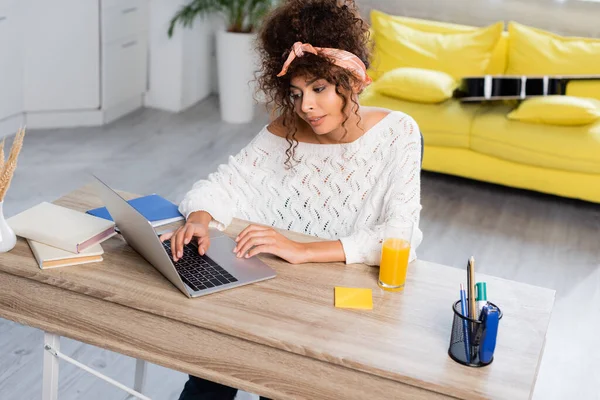 Curly freelancer looking at laptop near books and glass of orange juice on table — Stock Photo