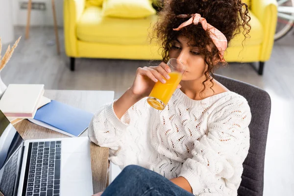 Кучерява жінка тримає скло і п'є апельсиновий сік біля ноутбука на столі — стокове фото