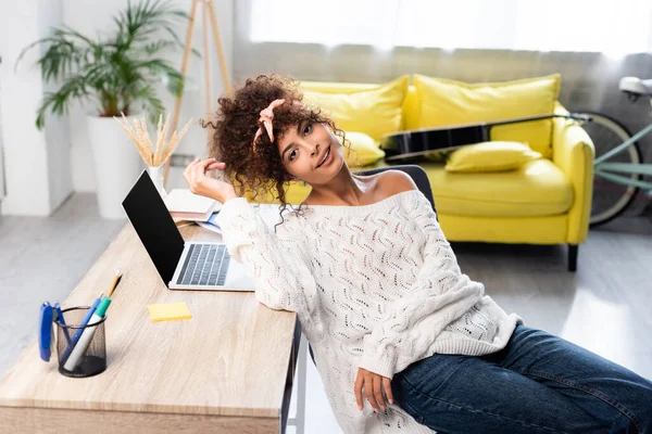 Mujer alegre sentada cerca del ordenador portátil con pantalla en blanco en el escritorio - foto de stock