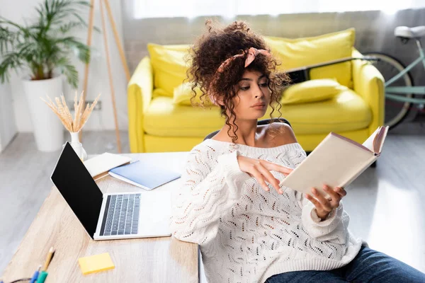 Bouclé femme lecture livre près d'un ordinateur portable avec écran blanc sur le bureau — Photo de stock