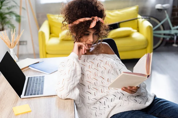 Jeune femme lecture livre près d'un ordinateur portable avec écran blanc sur le bureau — Photo de stock