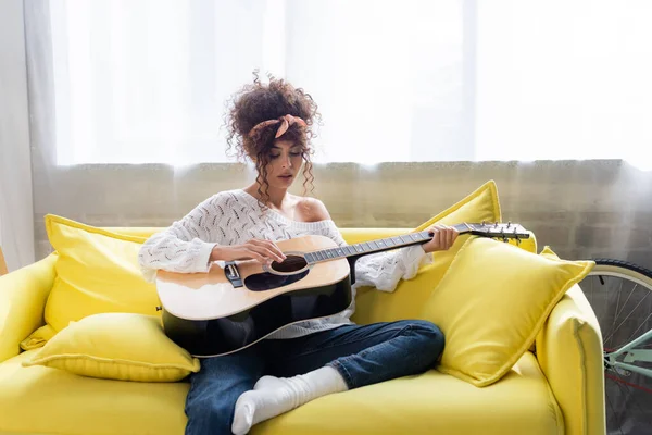 Mujer rizada tocando la guitarra acústica en el sofá en la sala de estar - foto de stock