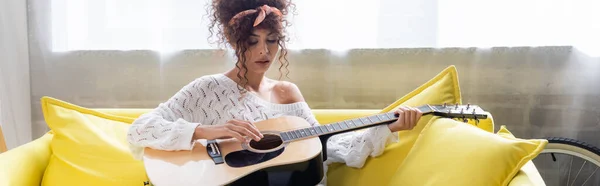 Panoramic shot of curly woman playing acoustic guitar on sofa in living room — Stock Photo