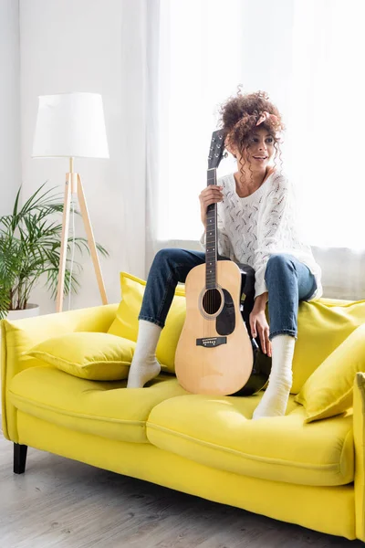 Довольная женщина с акустической гитарой и сидя на желтом диване в гостиной — стоковое фото