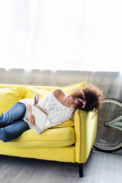 Молода жінка читає книгу, лежачи на дивані у вітальні — стокове фото