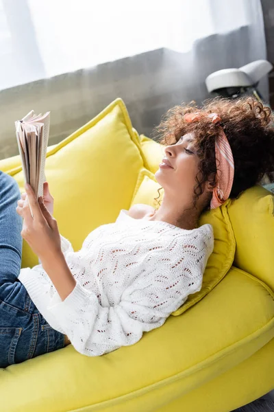 Mujer joven contenta leyendo libro mientras está acostado en el sofá en la sala de estar - foto de stock